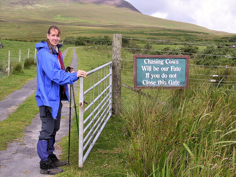 Woman standing next to a sign which reads Chasing Cows will be our fate if you don't close this gate