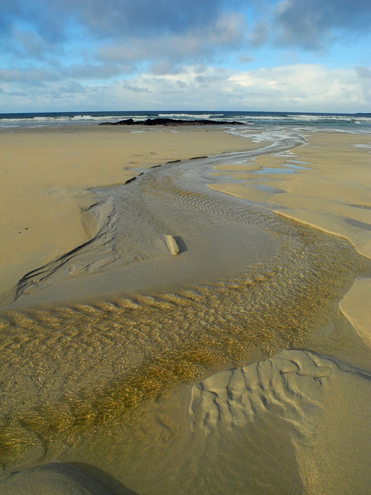 Picture of a small burn with slightly peaty brown water running over a beach