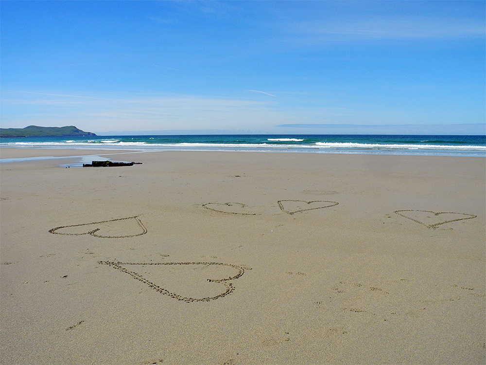 Picture of a beach in the sunshine with a number of hearts drawn in the sand