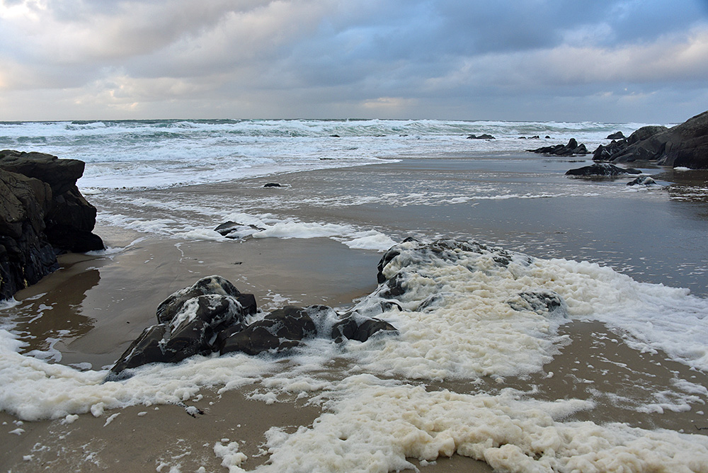 Picture of a beach with rocks, foam around the rocks, waves in the background