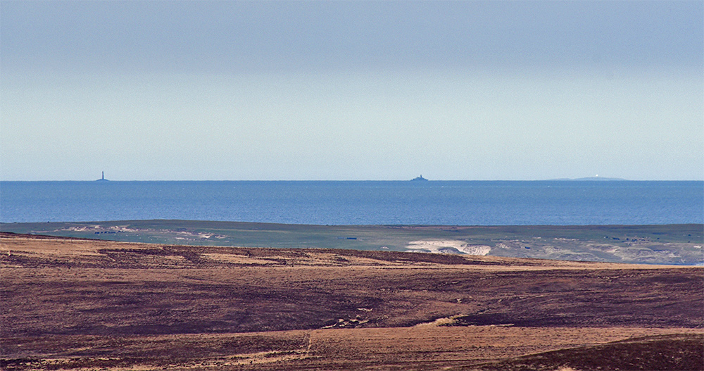 Picture of a view out to sea from a hill, a lighthouse and a warship in the distance