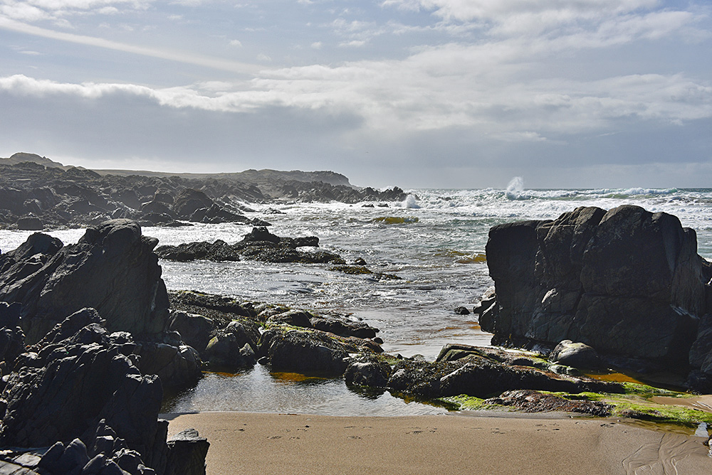 Picture of a view out to sea between two rocks and some sandy beach on a breezy day