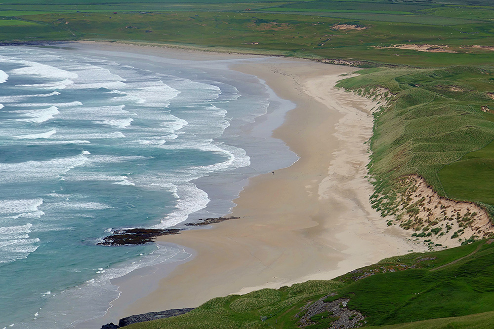 Picture of a view from a hill down to a sandy beach with a lone walker on it