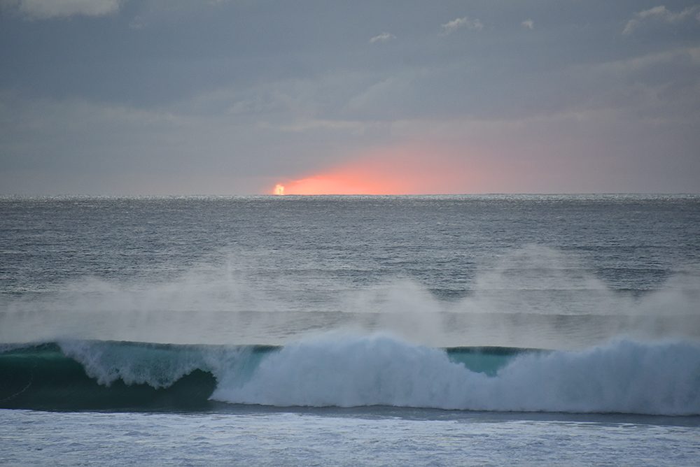 Picture of a cloudy sunset with a tiny glimpse of the red sun on the horizon, waves breaking in the foreground with spray in the air