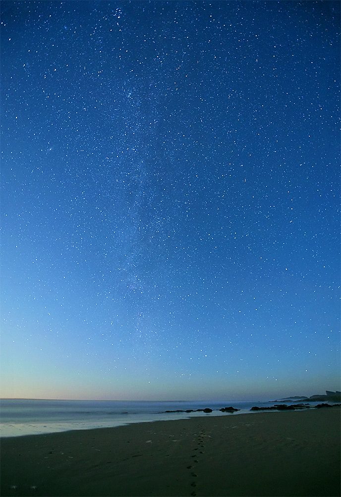 Picture of a starry night sky with the Milky Way over a beach with a line of footprints in the sand