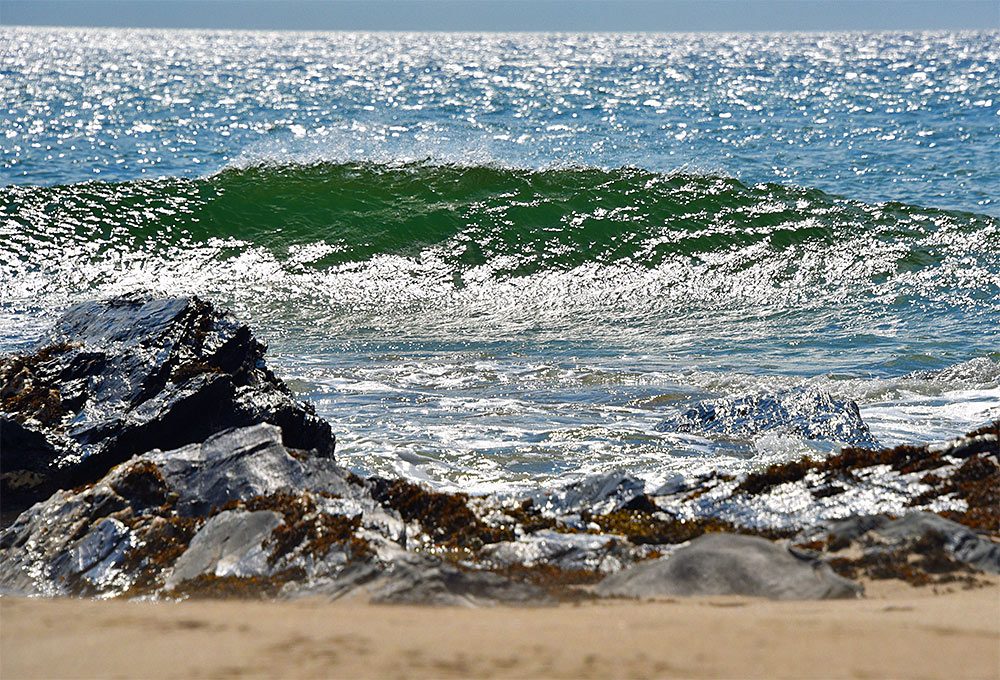 Picture of bright sunlight reflecting on water, a wave breaking, seen from a beach with some rocks