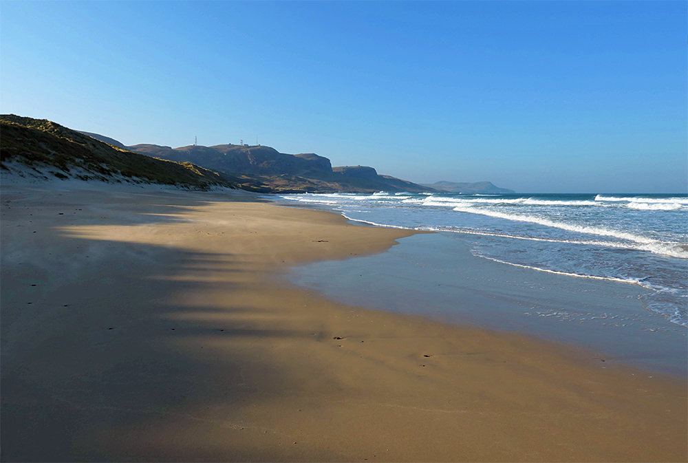 Picture of a sandy beach in a wide bay with waves rolling in, shades from the dunes covering half the beach