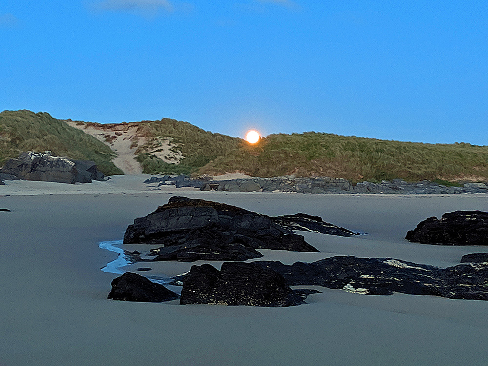 Picture of the Moon rising over dunes behind a beach with some rocks