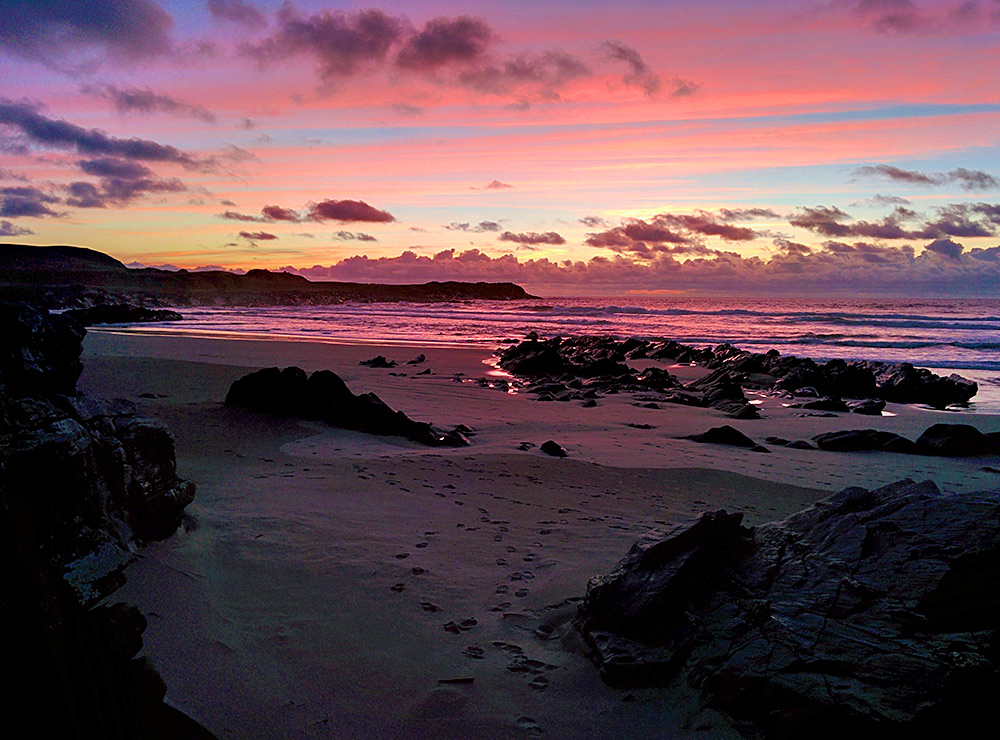 Picture of a colourful sky mirrored on the water of a bay with a sandy beach and some rocks