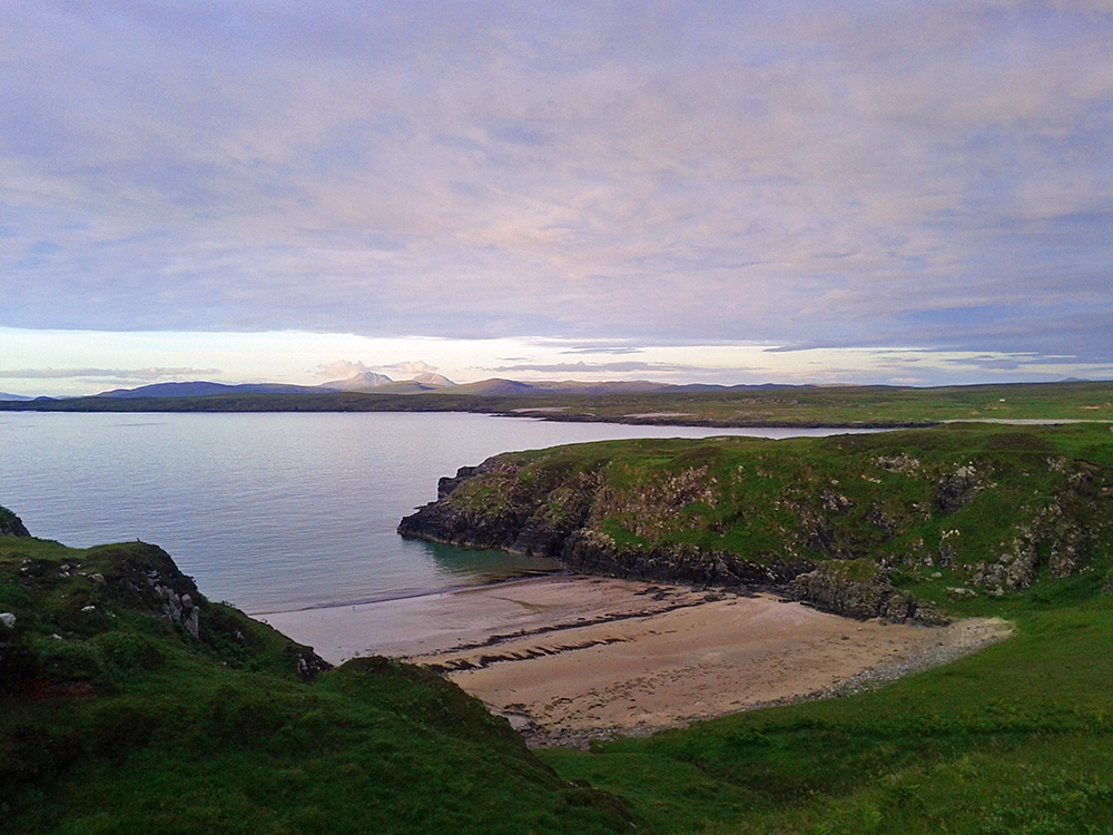 Picture of a small beach between cliffs, a bay behind and mountains in the distance