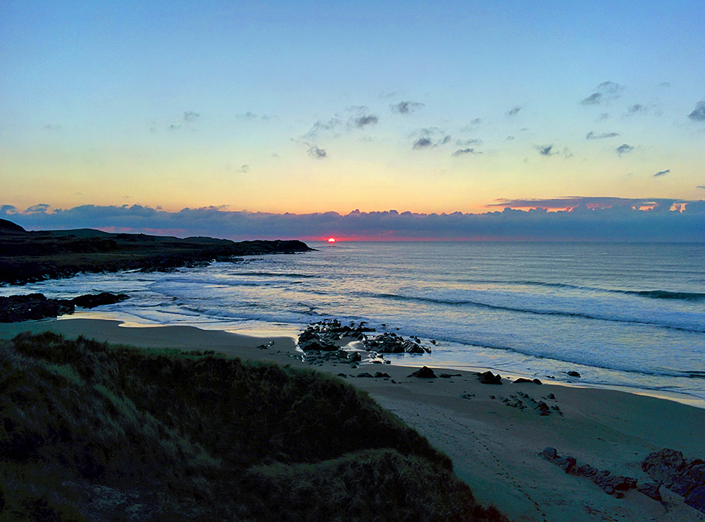 Picture of the last seconds of a sunset watched from the dunes behind a bay with a beach, the sun half behind the horizon