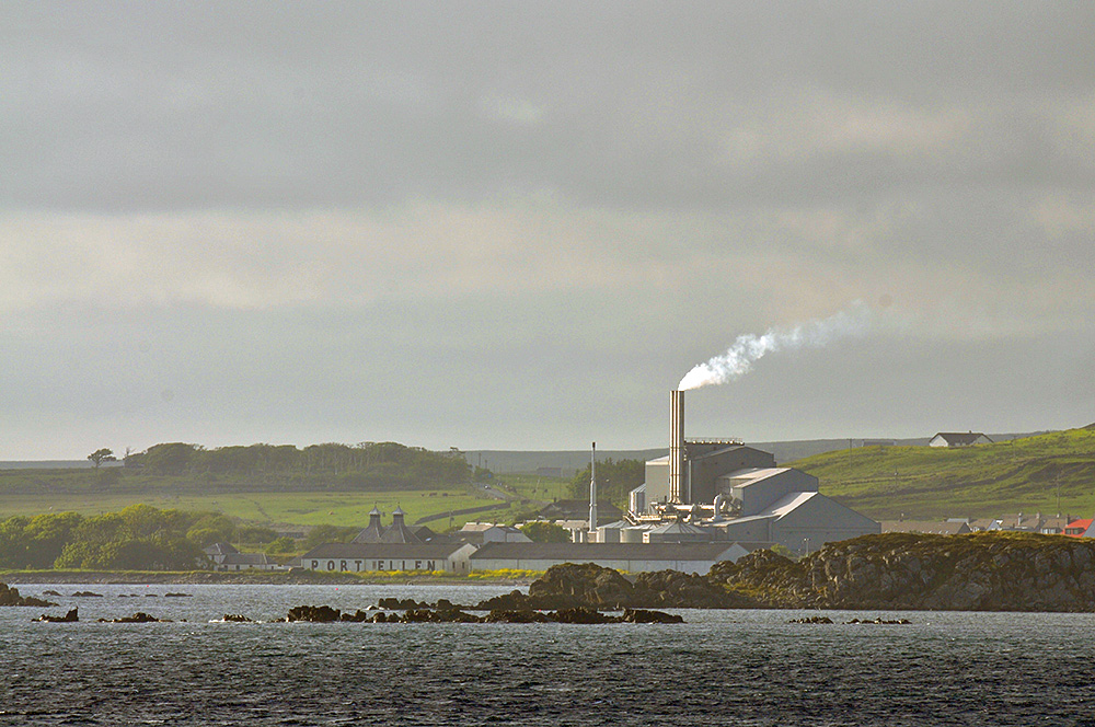 Picture of a coastal distillery and maltings behind some skerries in mild summer evening light
