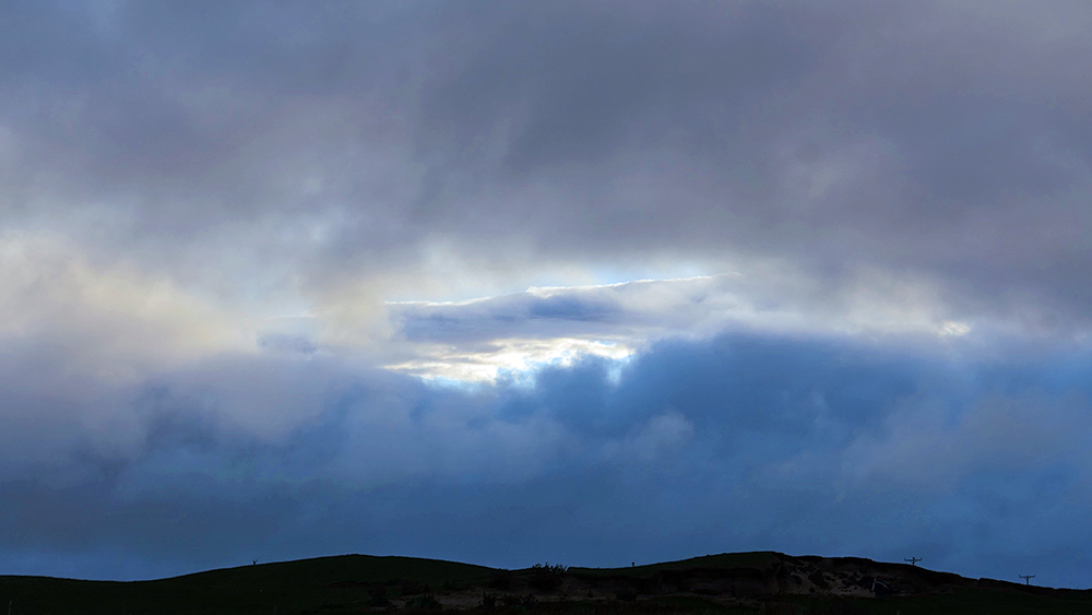 Picture of a break in dark clouds with some other clouds behind it, seen over some dunes