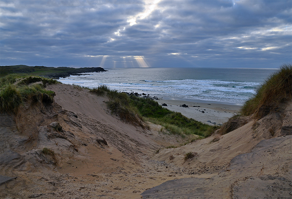 Picture of sun rays breaking through clouds over a coast with rocks and beach, seen from/through dunes