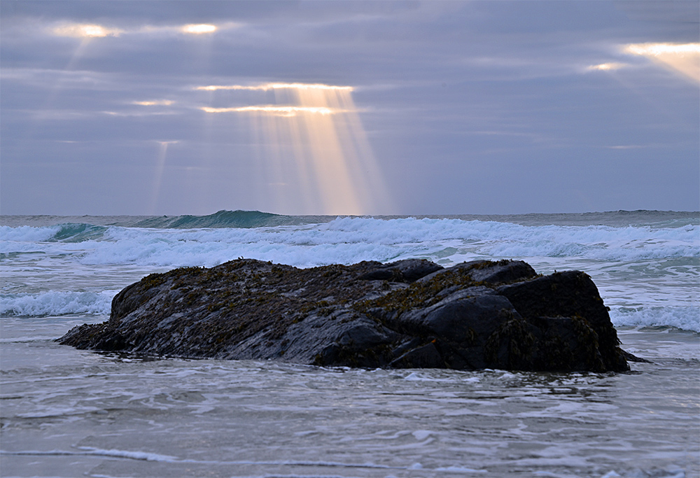 Picture of sun rays through the clouds and waves behind a big rock on a beach