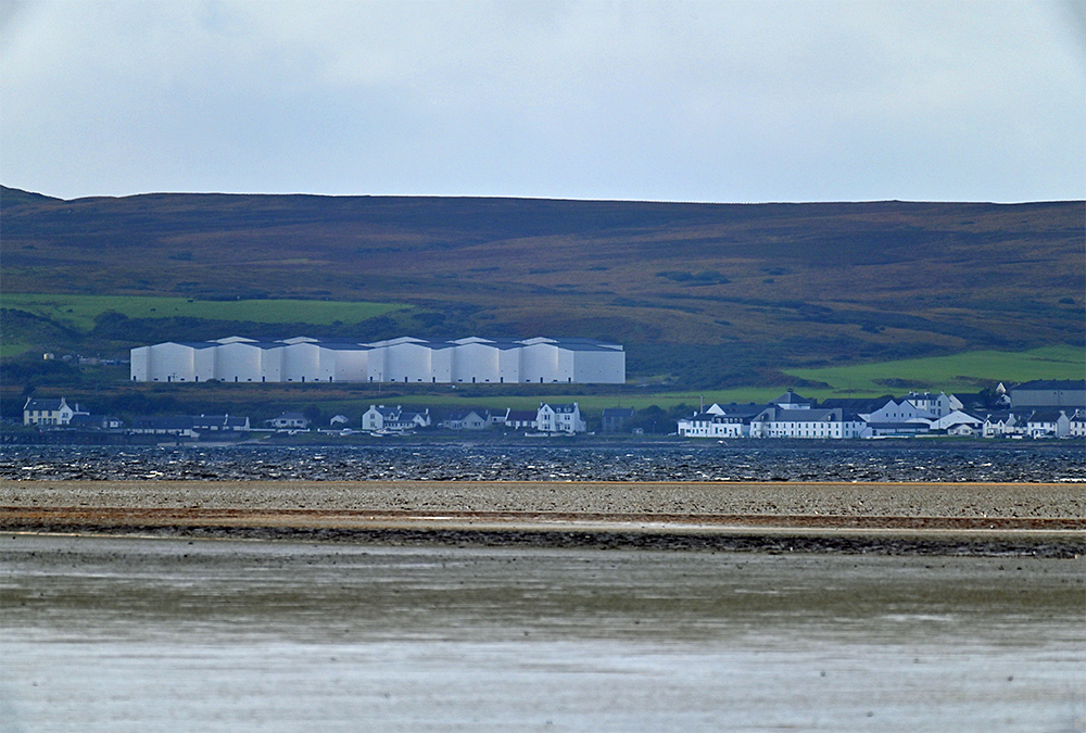Picture of distillery warehouses above a coastal village, seen across a sea loch