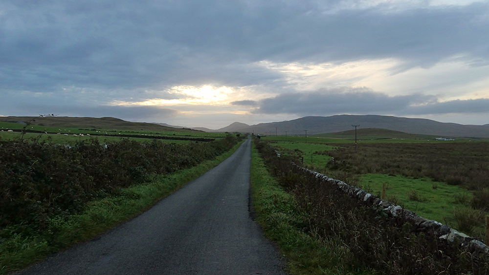 Picture of a single track road on a mostly overcast morning with some brightness around the rising sun