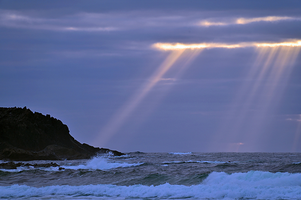 Picture of sun rays breaking through clouds over a coast with rocks and breaking waves