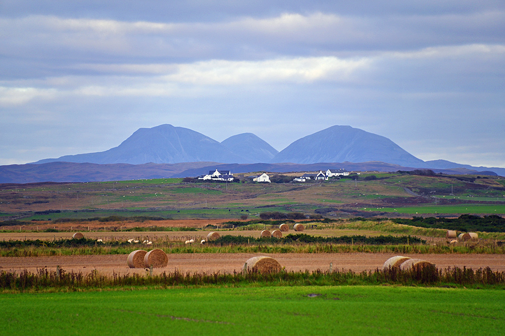 Picture of three mountains behind a small settlement and some fields with hay bales