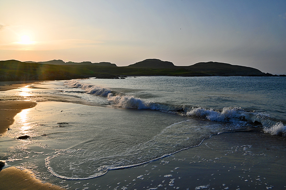 Picture of a low late afternoon sun over hills behind a bay with a sandy beach, a wave breaking on the beach