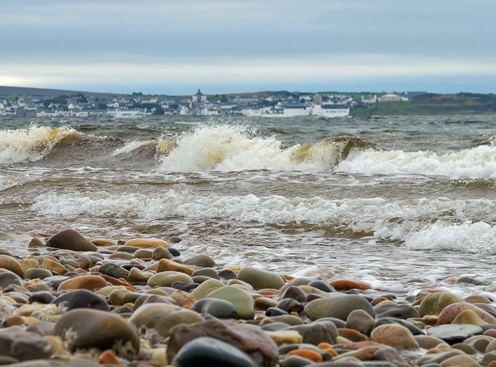 Picture of pebble beach with waves rolling in, a coastal village in the background on the other side of the loch