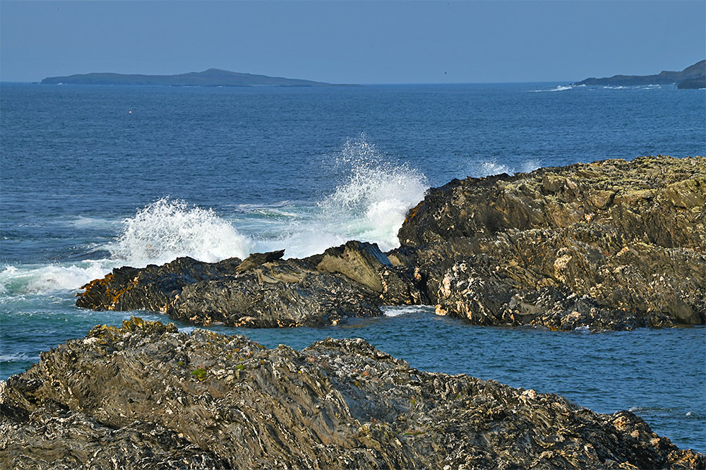 Picture of a wave breaking and splashing at some coastal rocks, a small island in the background