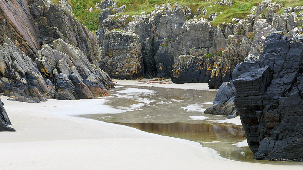 Picture of a sandy beach gap between rocks, a shallow burn flowing through the gap