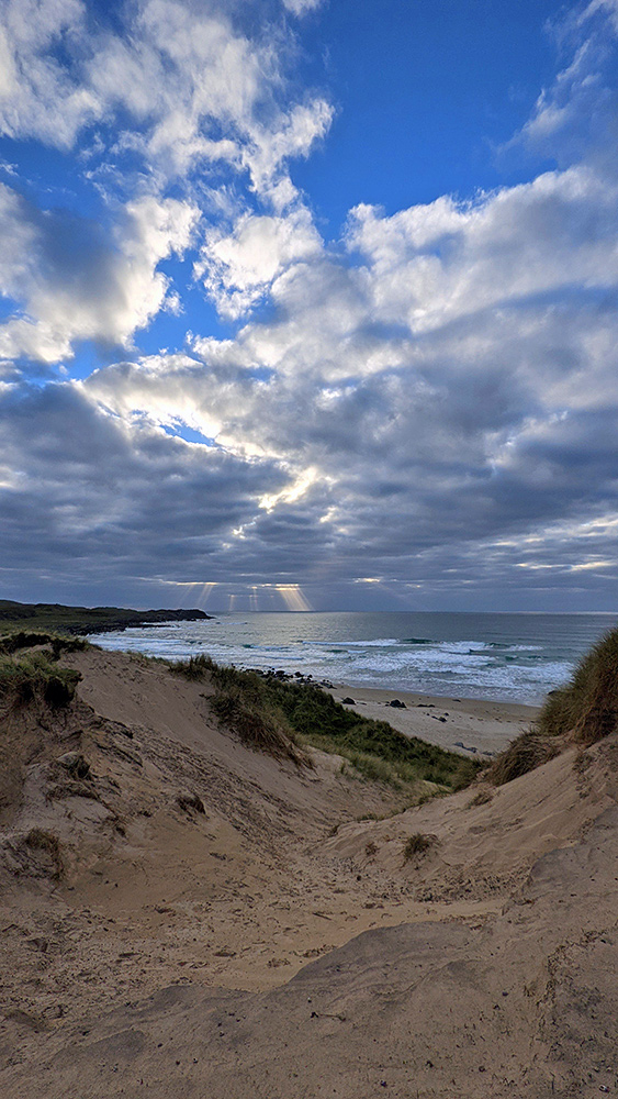 Picture of some distant sunrays breaking through clouds seen from the top of dunes over a bay with a beach