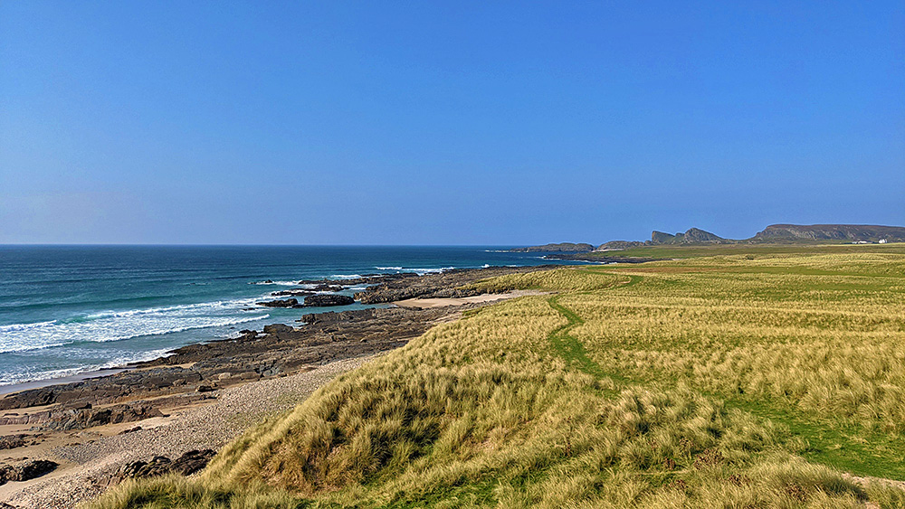 Picture of a view along a coastline with dunes, beach, rocks and cliffs. A track leading along the dunes into the distance