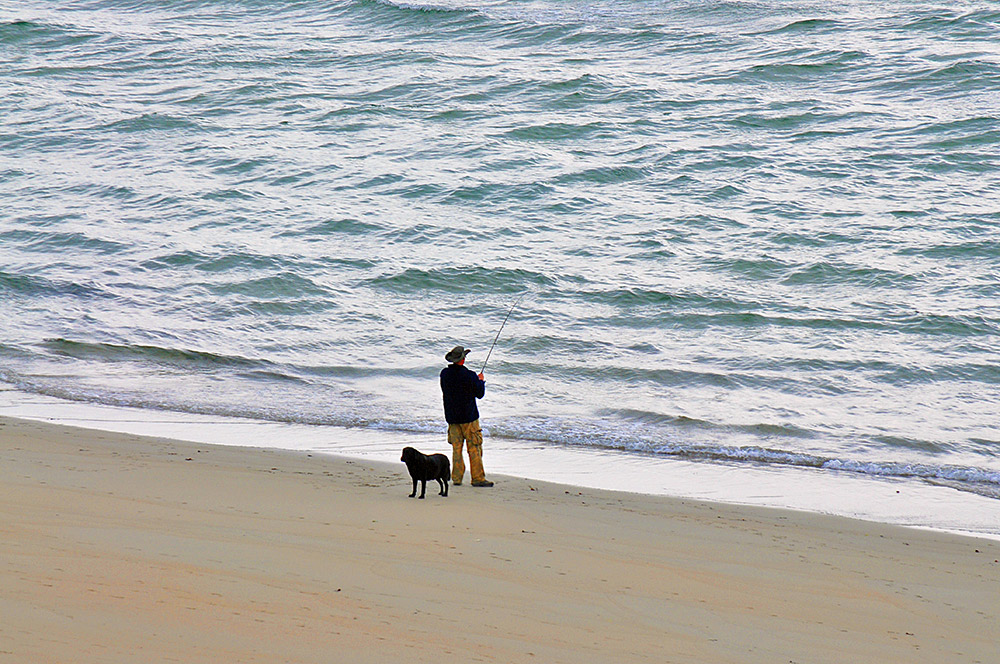 Picture of a man angling on a beach with his dog standing behind him