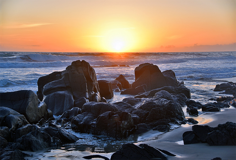 Picture of rocks on a beach with waves rolling in behind during a summer sunset