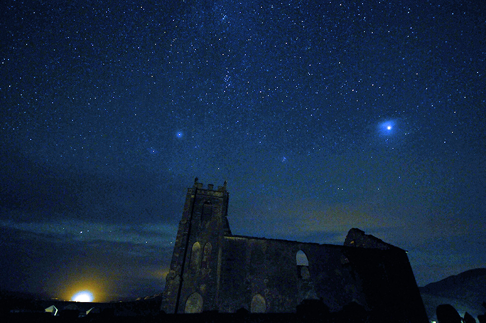 Picture of a starry night sky above the ruin of a church, a bright light in the distance on the side