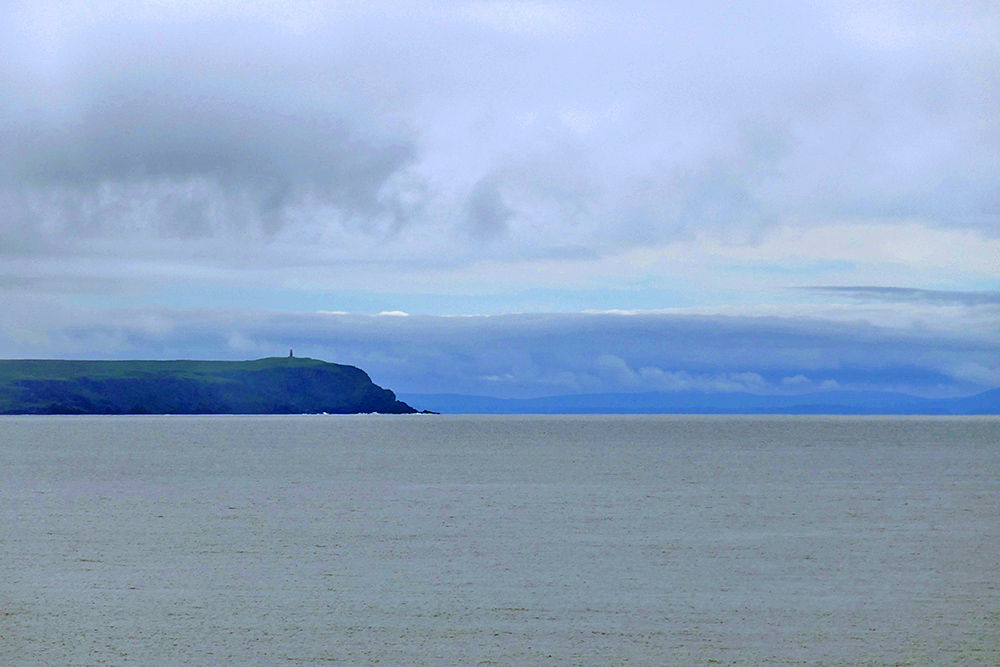 Picture of a mull with steep cliffs seen across the sea, another coastline visible in the distance
