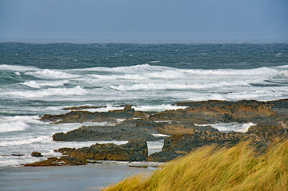 Picture of big waves rolling into a bay with a shore with a mixture of rocks and beach below some dunes