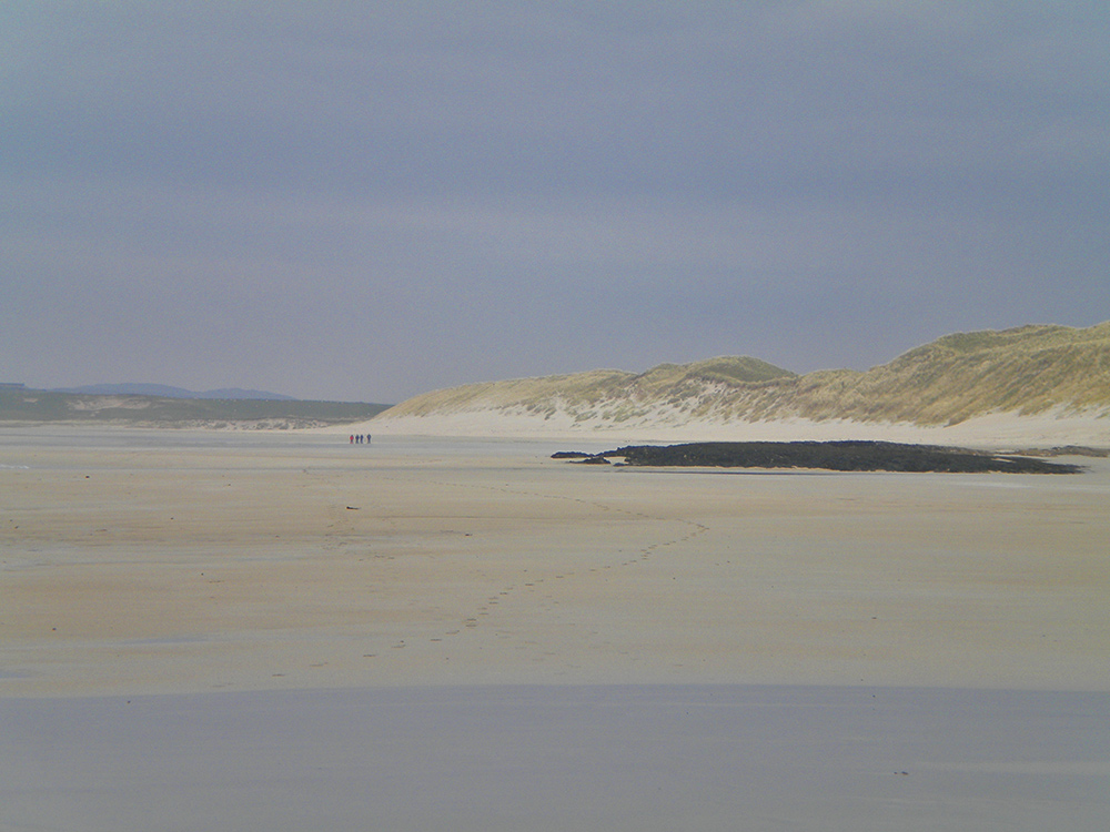 Picture of four distant walkers on a wide beach with dunes behind on a hazy day