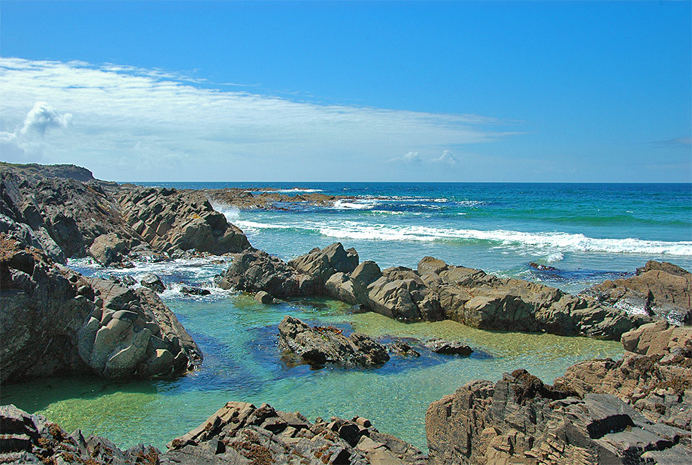 Picture of a rocky shoreline with a rockpool at a bay, waves rolling in on a sunny day