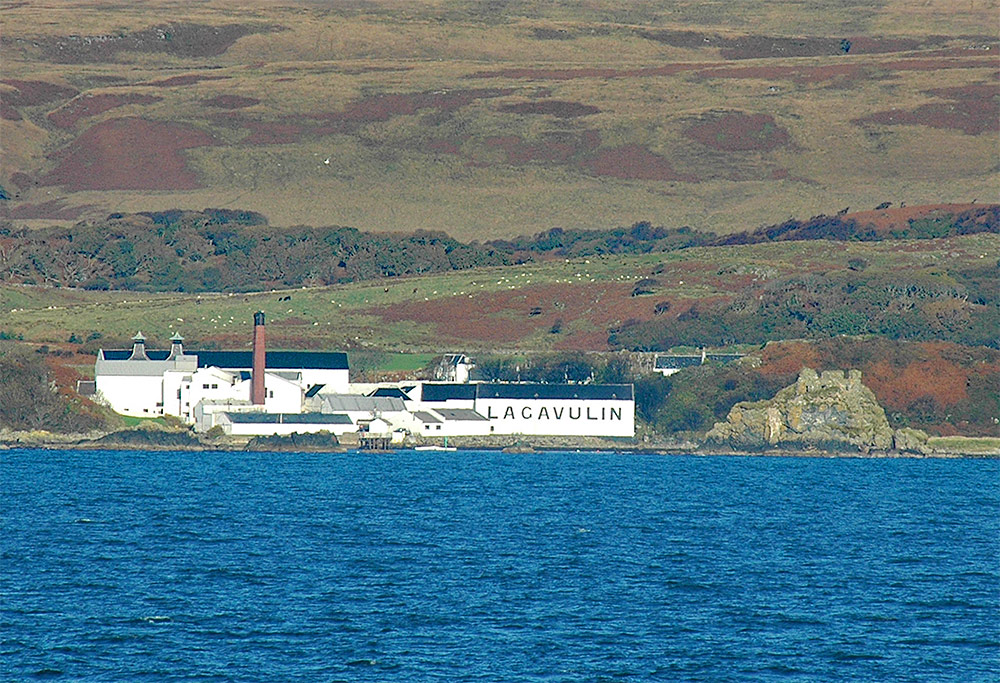 Picture of a coastal distillery (Lagavulin on Islay) with an old castle ruin near it, seen from a passing ferry