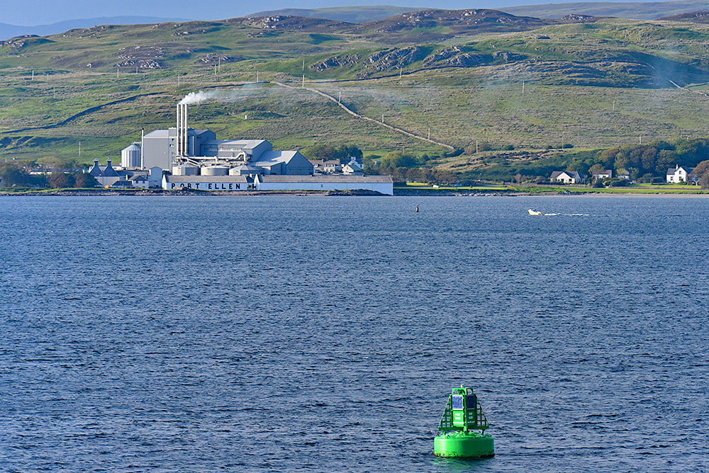 Picture of a green buoy in a bay with a distillery and maltings on the shore. Also a motorboat passing.