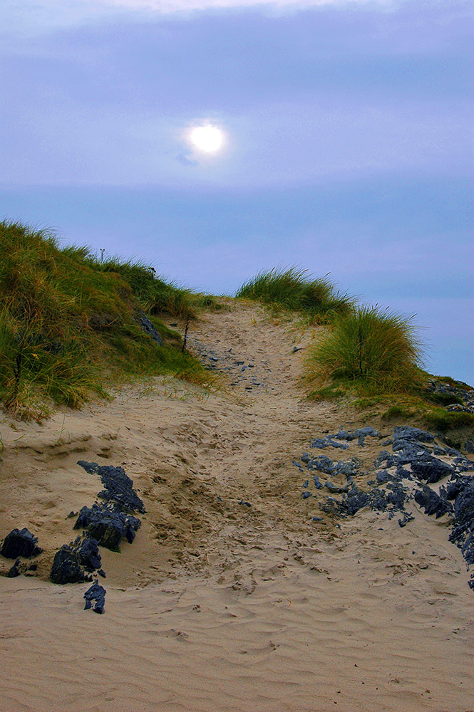 Picture of a path leading up some dunes, hazy sunshine breaking through some clouds above