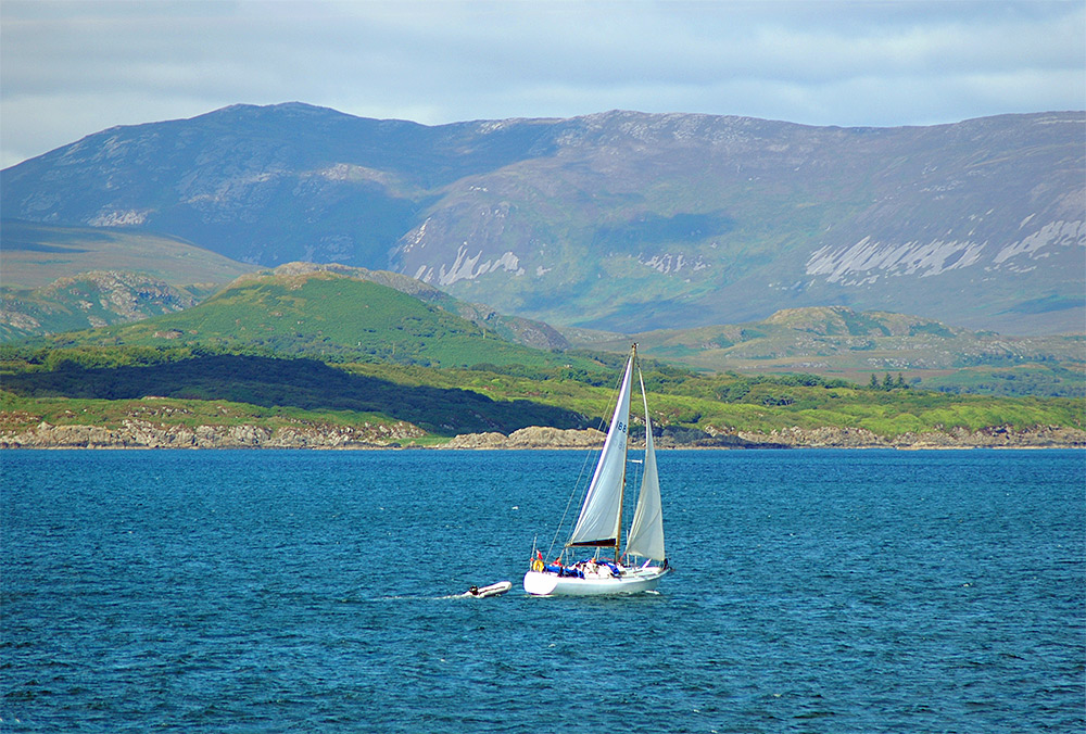Picture of a sailing yacht passing a coastline with a small mountain and hills