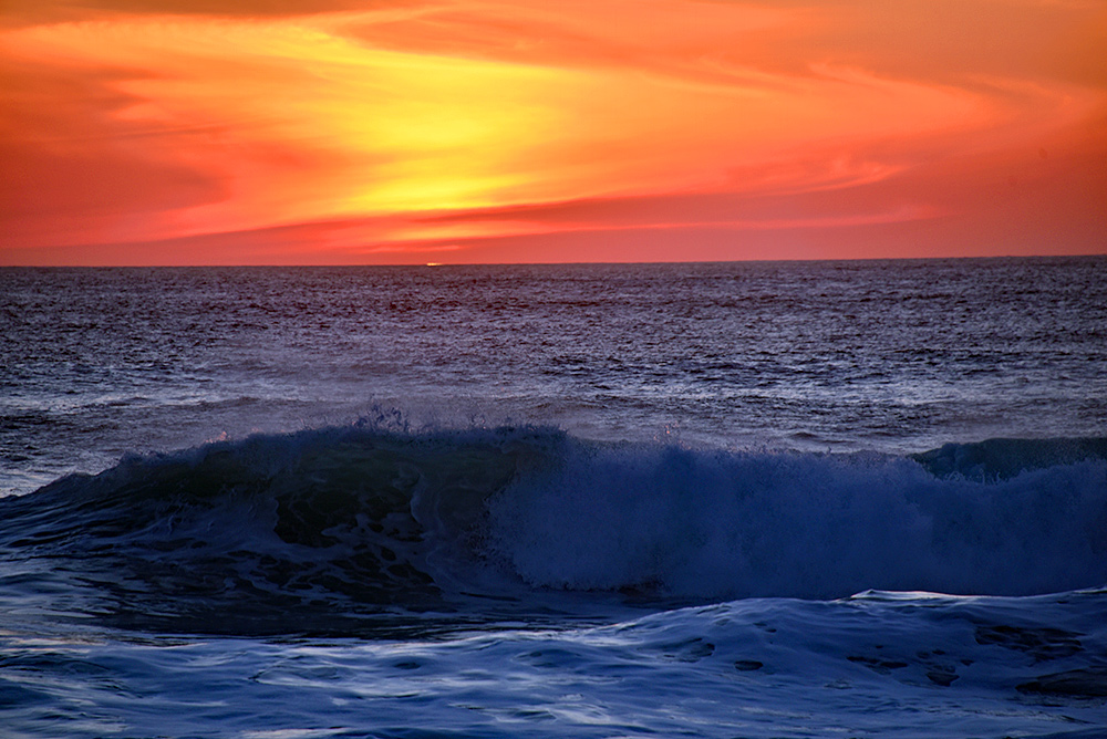 Picture of a wave breaking as it reaches a shore, a bright orange and red sky at sunset in the background