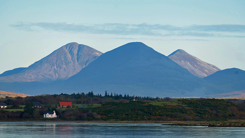 Picture of three mountains seen across a sea loch, the front middle one in the dark, two distant ones behind it in the light