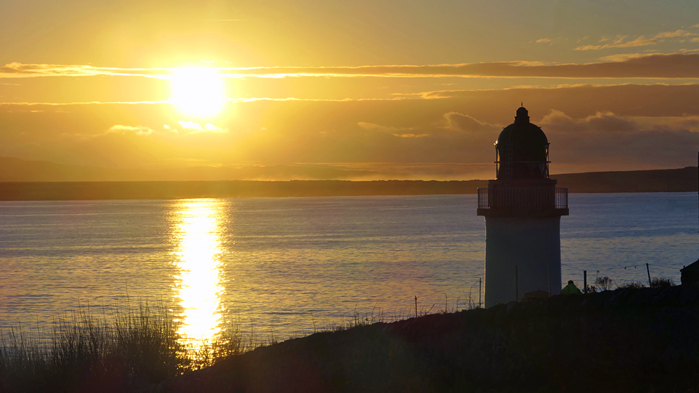 Picture of a small lighthouse at a sea loch with the sun rising above the loch