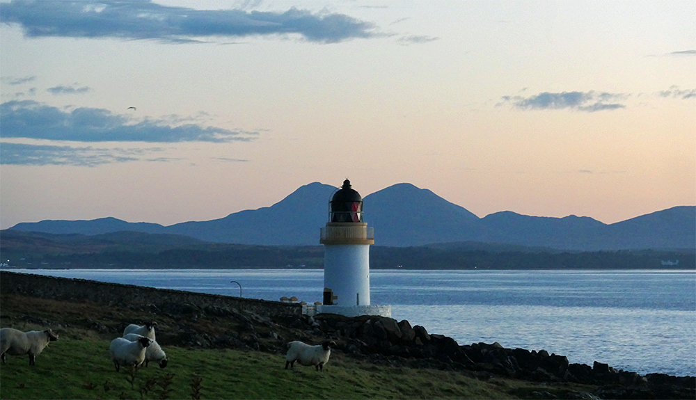 Picture of small lighthouse at dawn, some sheep grazing in front, some mountains in the distance