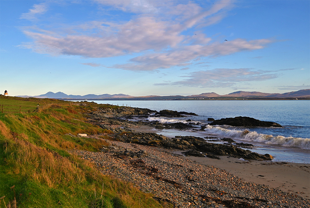 Picture of a calm sea loch seen from a small beach near a village, the top of a lighthouse just visible behind a wall
