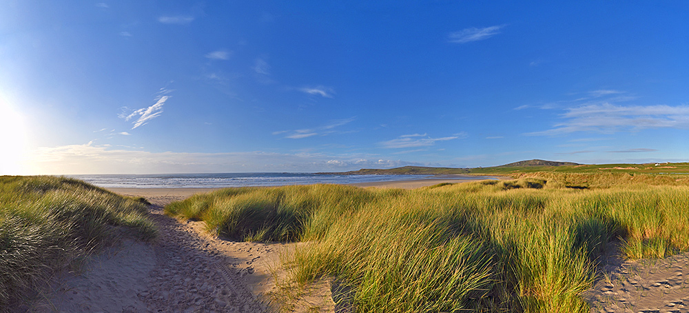 Panoramic picture of the view from a path through dunes to a bay with a wide sandy beach