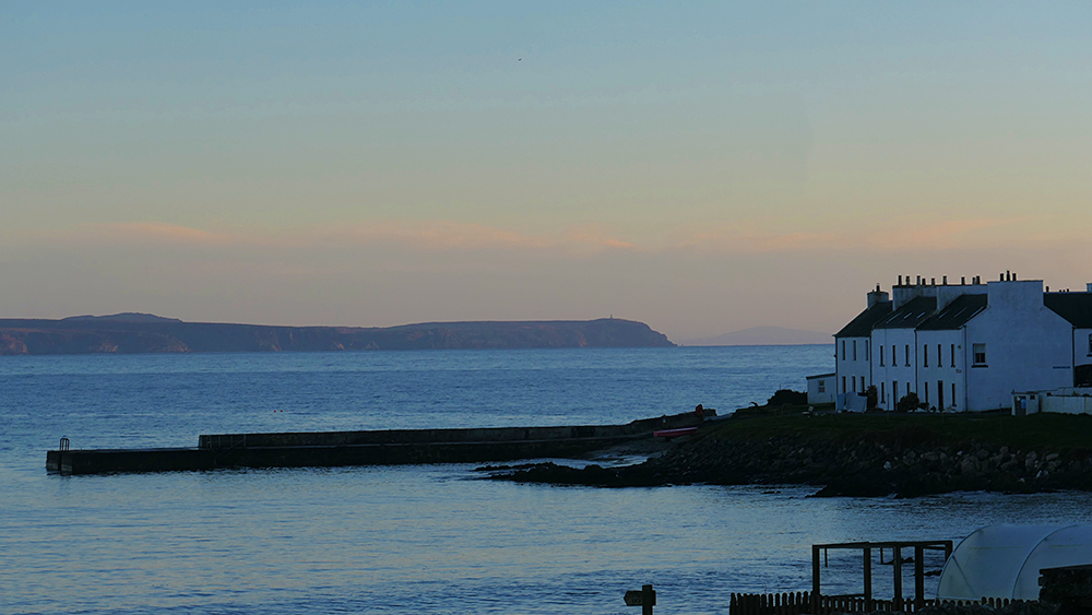 Picture of coastal village with a pier, a peninsula across a sea loch in the distance in the mild dusk light