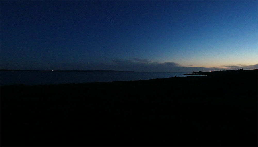 Picture of a dark coastal landscape in the very last light, a village just visible in the distance
