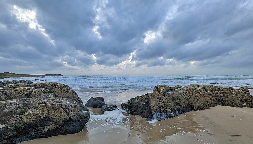 Picture of a beach with some large rocks under some clouds with waves rolling into the bay
