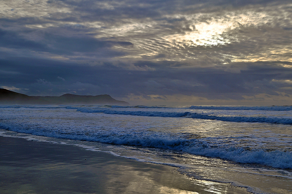 Picture of waves rolling on to a beach in a bay under some heavy clouds with some sun rays breaking through